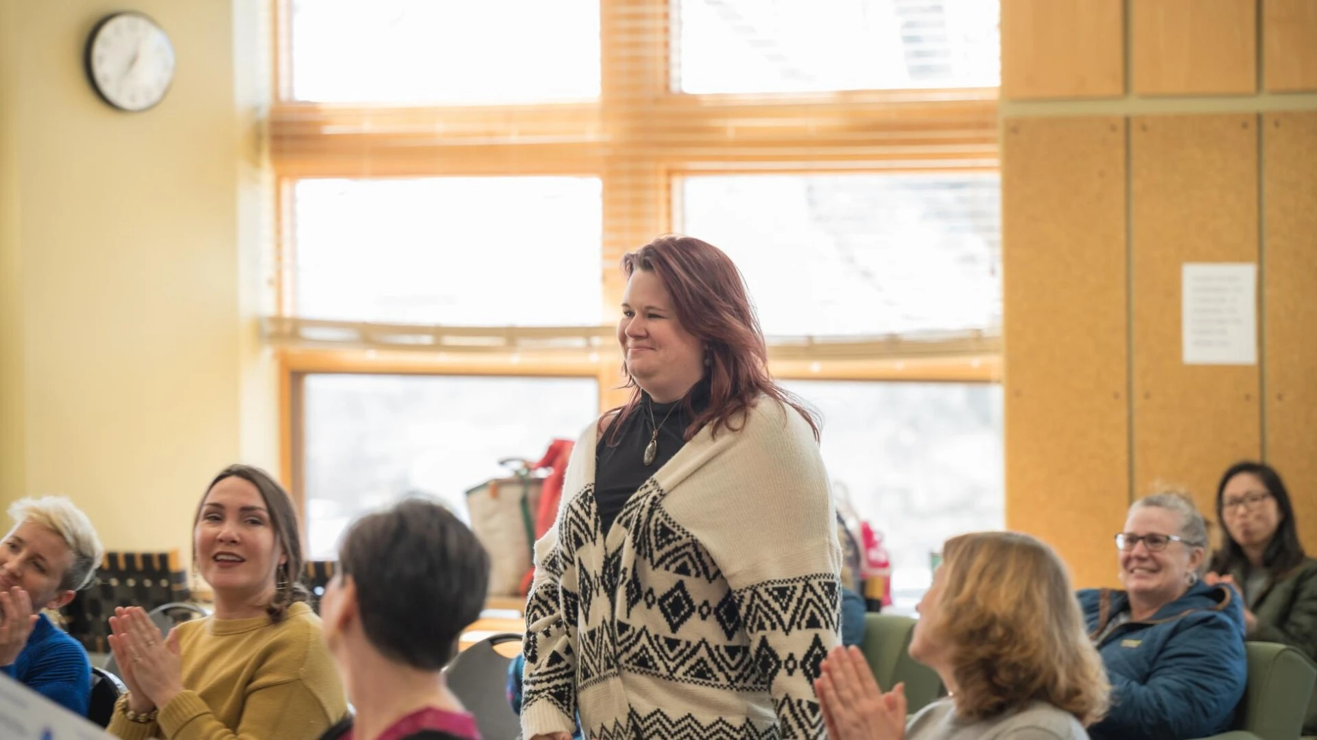 A person standing in the middle of a room smiling, as people sitting around them clap and smile at them.