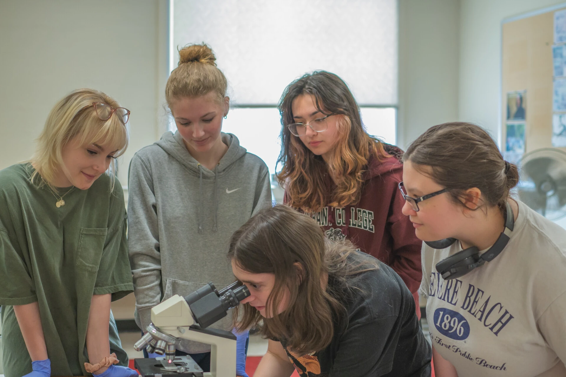 Students in a biology classroom setting with one female student looking into a microscope with other female students watching