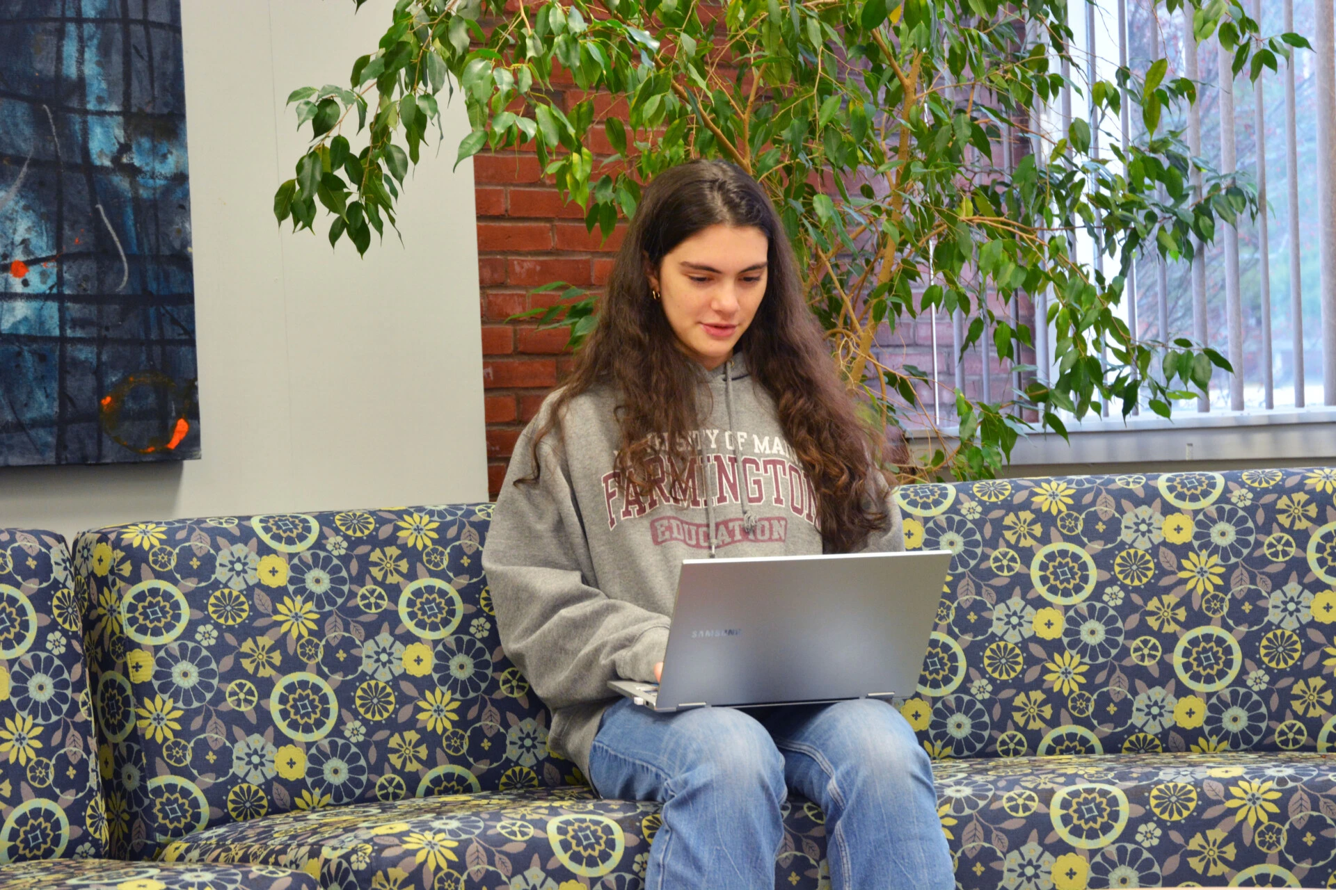 Female student wearing a University of Maine Farmington sweater attentively using her laptop while sitting on a wide couch.