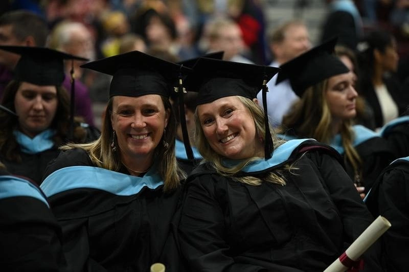 Two graduate students sitting smiling at the camera during UMF commencement with their regalia on.
