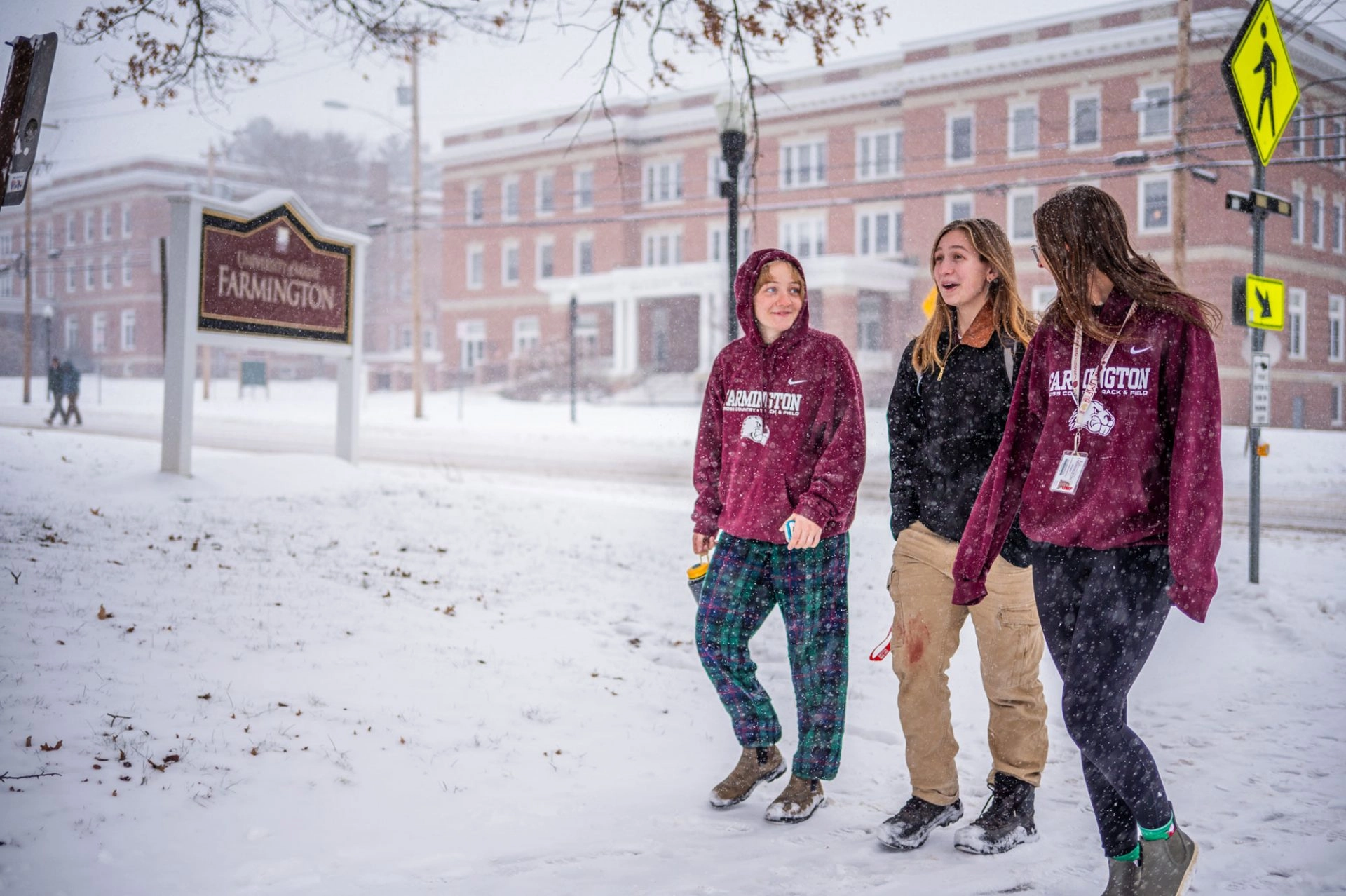 Three students walking through snow flakes in winter on a path in front of a University of Maine Farmington sign and building.