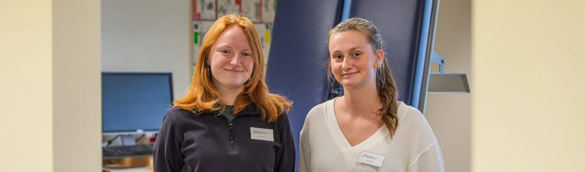 two chiropractic student interns stand side by side in from of the chiropractic bed in the office.