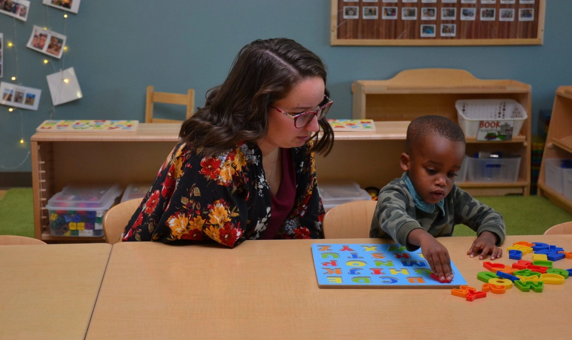 Teacher sitting with young student looking at alphabet board on table