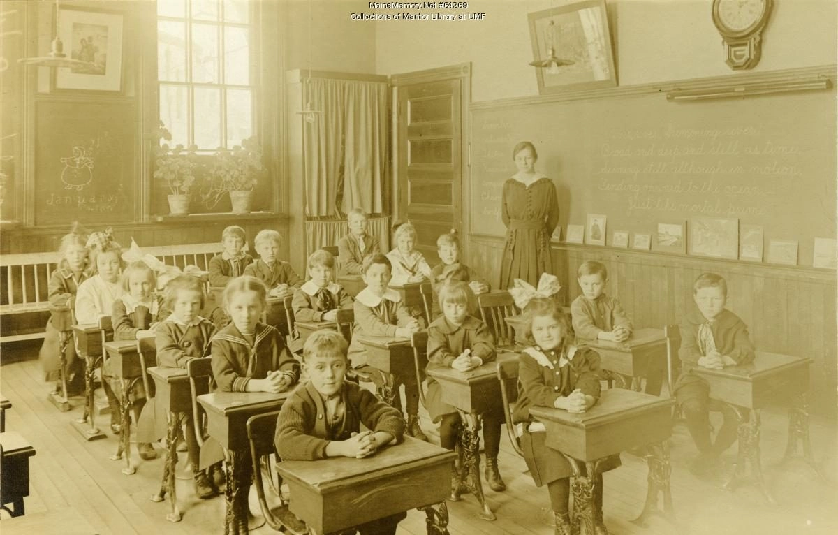 Black and white image of model classroom from 1917 with students at a desk and teacher in the back