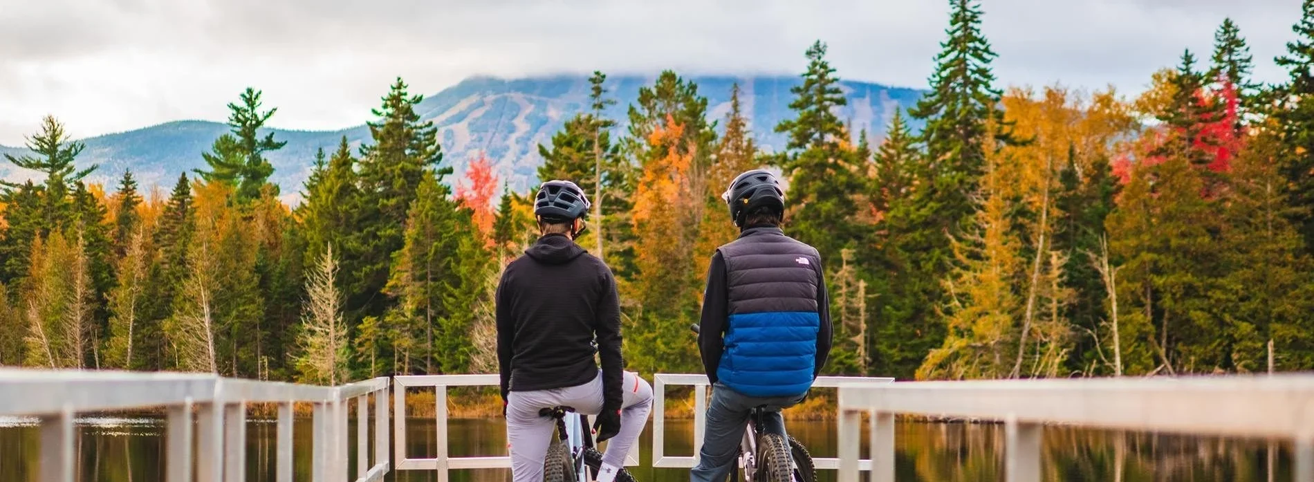 Two bikers on a dock in front of a lake and fall colors with a mountain in the distance