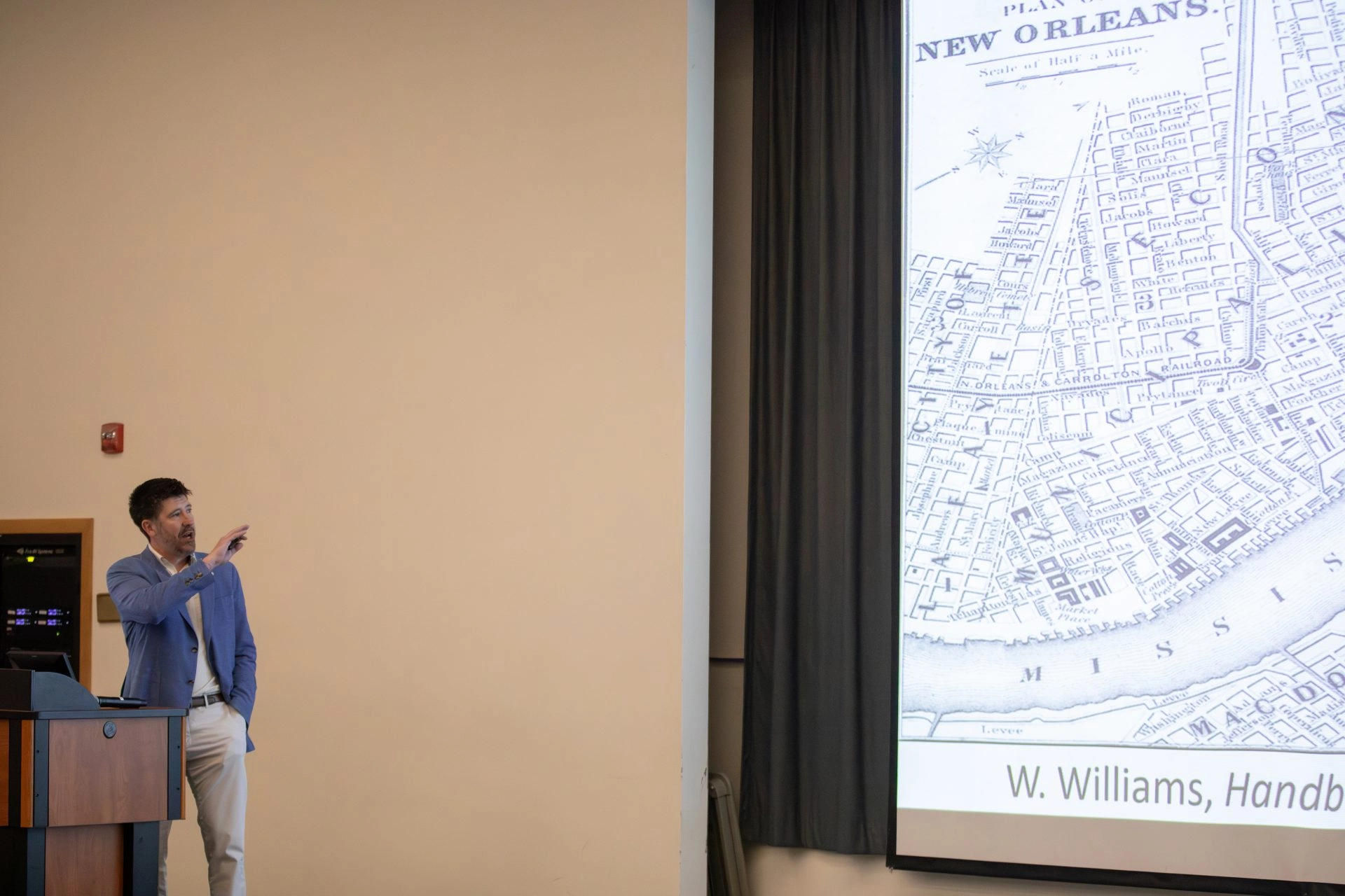 Professor standing in front of auditorium pointing at a projection of a map of New Orleans