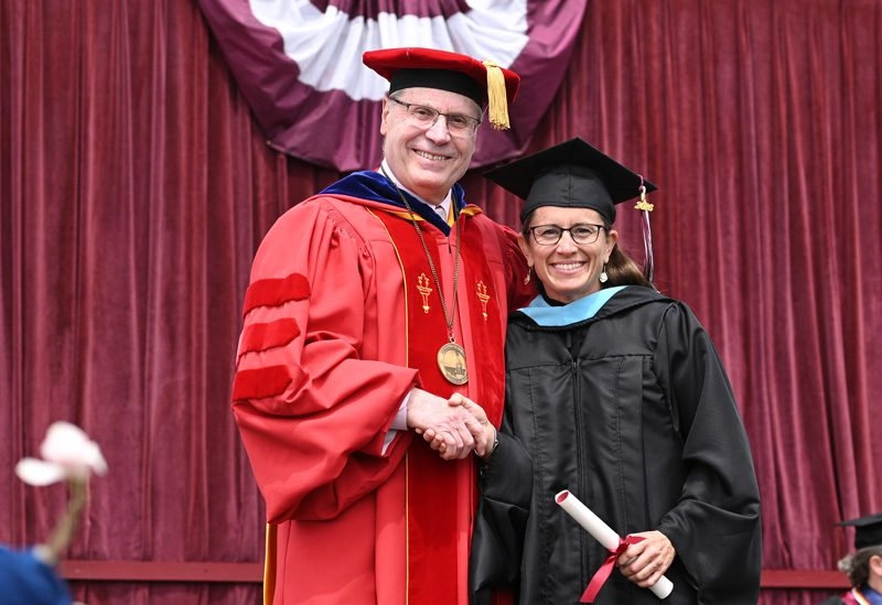 Student shaking hand of UMF president holding degree on commencement stage smiling