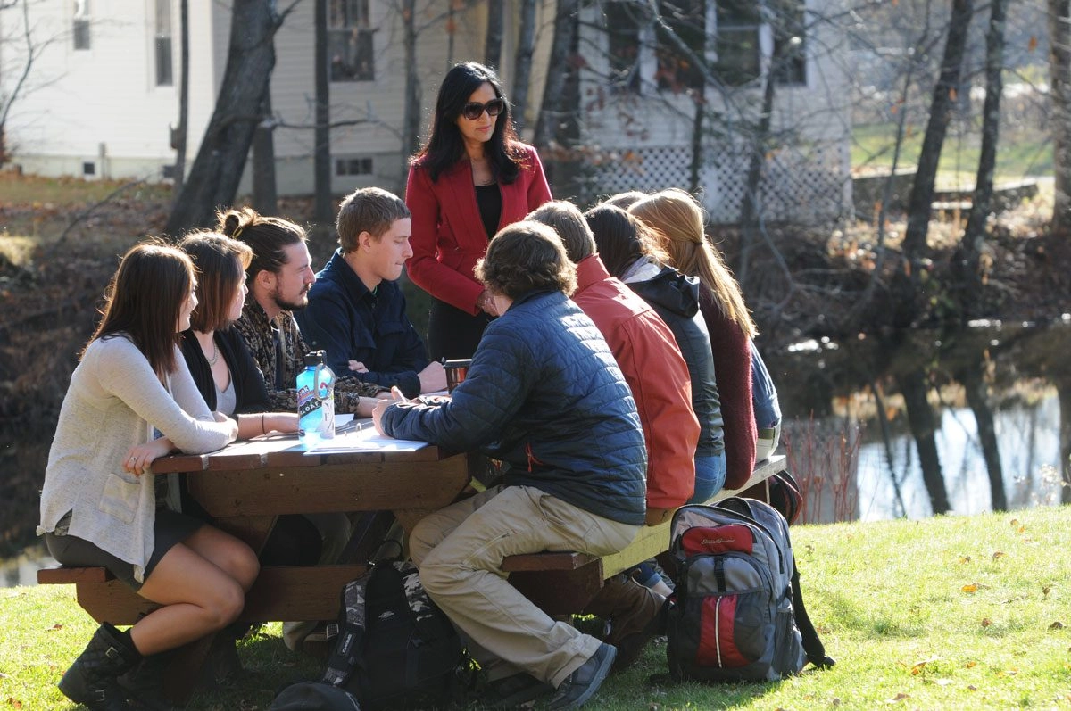 Group of students sitting at a picnic table outside in front of a bond listening to a professor standing in front of the table.
