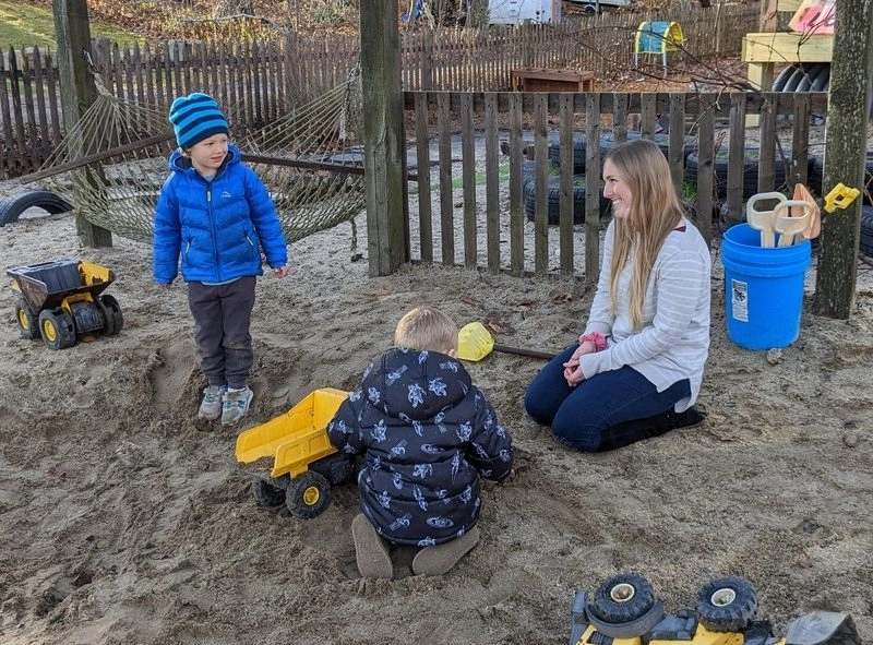 Teacher outside in playground sandbox with two young boys playing with trucks
