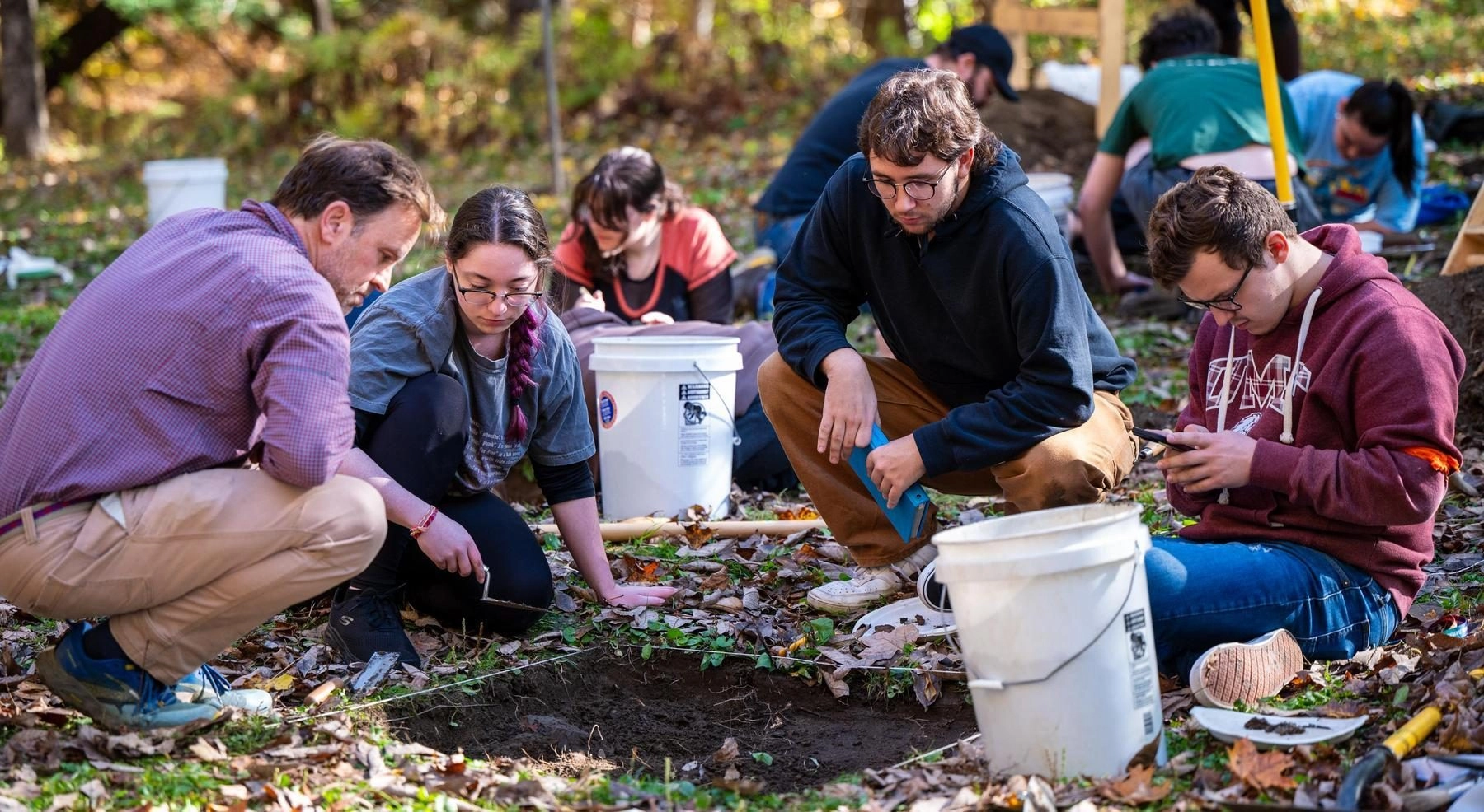 Group of young adults excavating soil in a park, kneeling with trowels and buckets nearby.