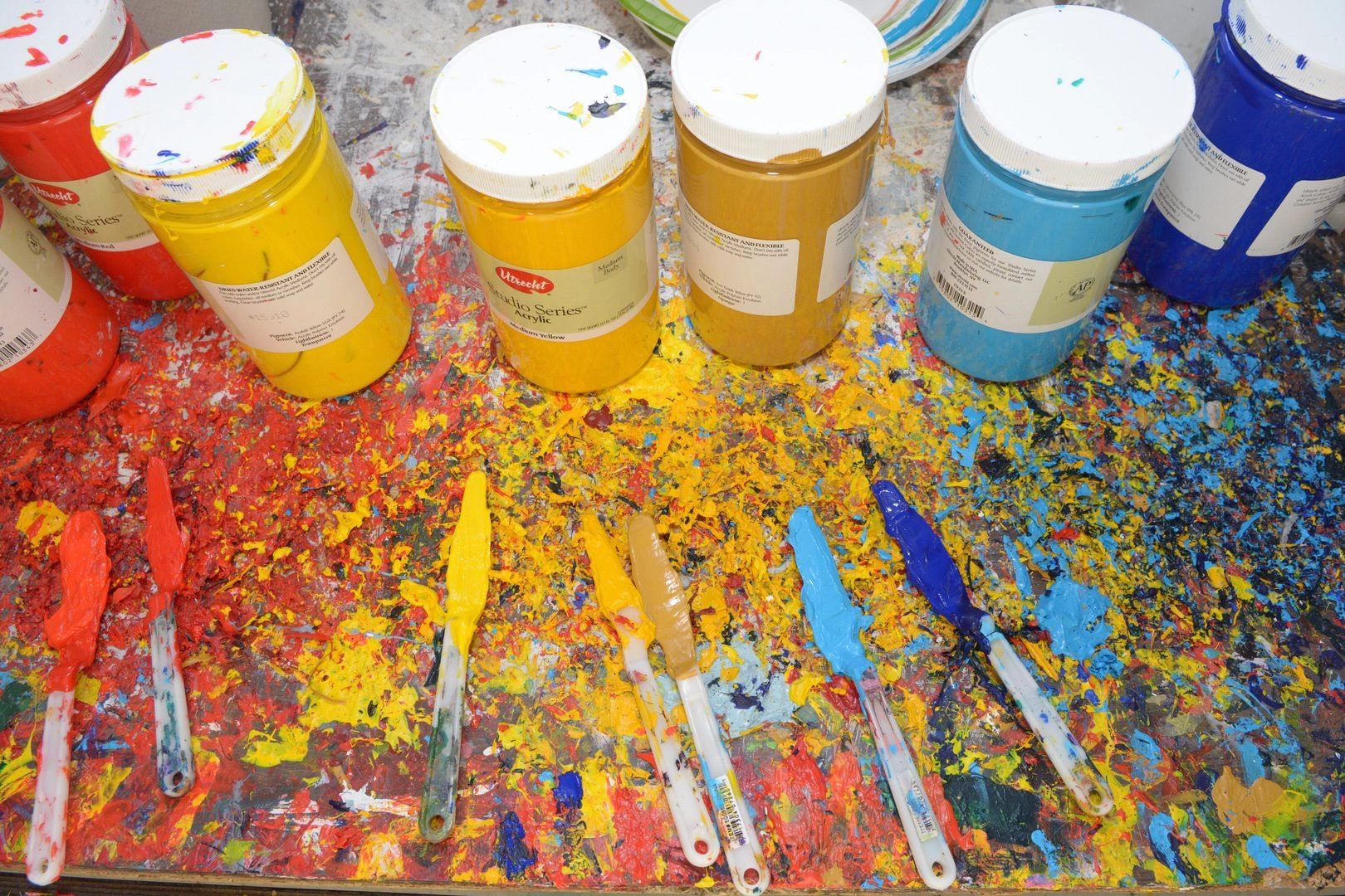 Row of paint jars in red, yellow, orange, and blue on a splattered art table with paint-smeared brushes.