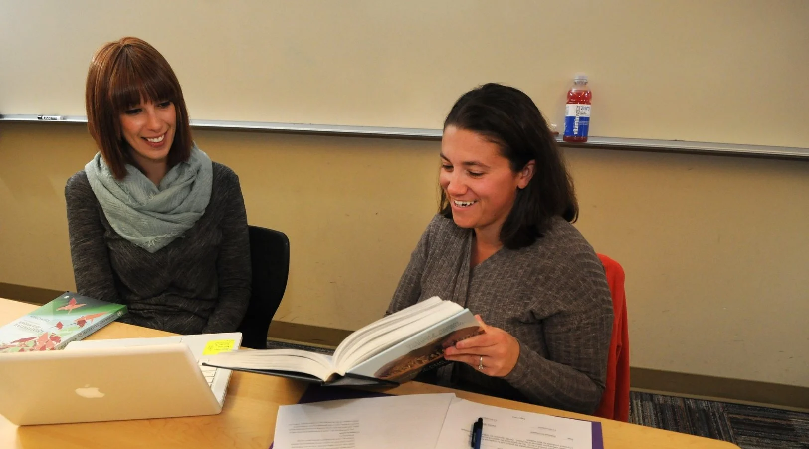 Two women sit at a table in a classroom, one smiles while reading a thick book; a laptop and papers are on the desk nearby.