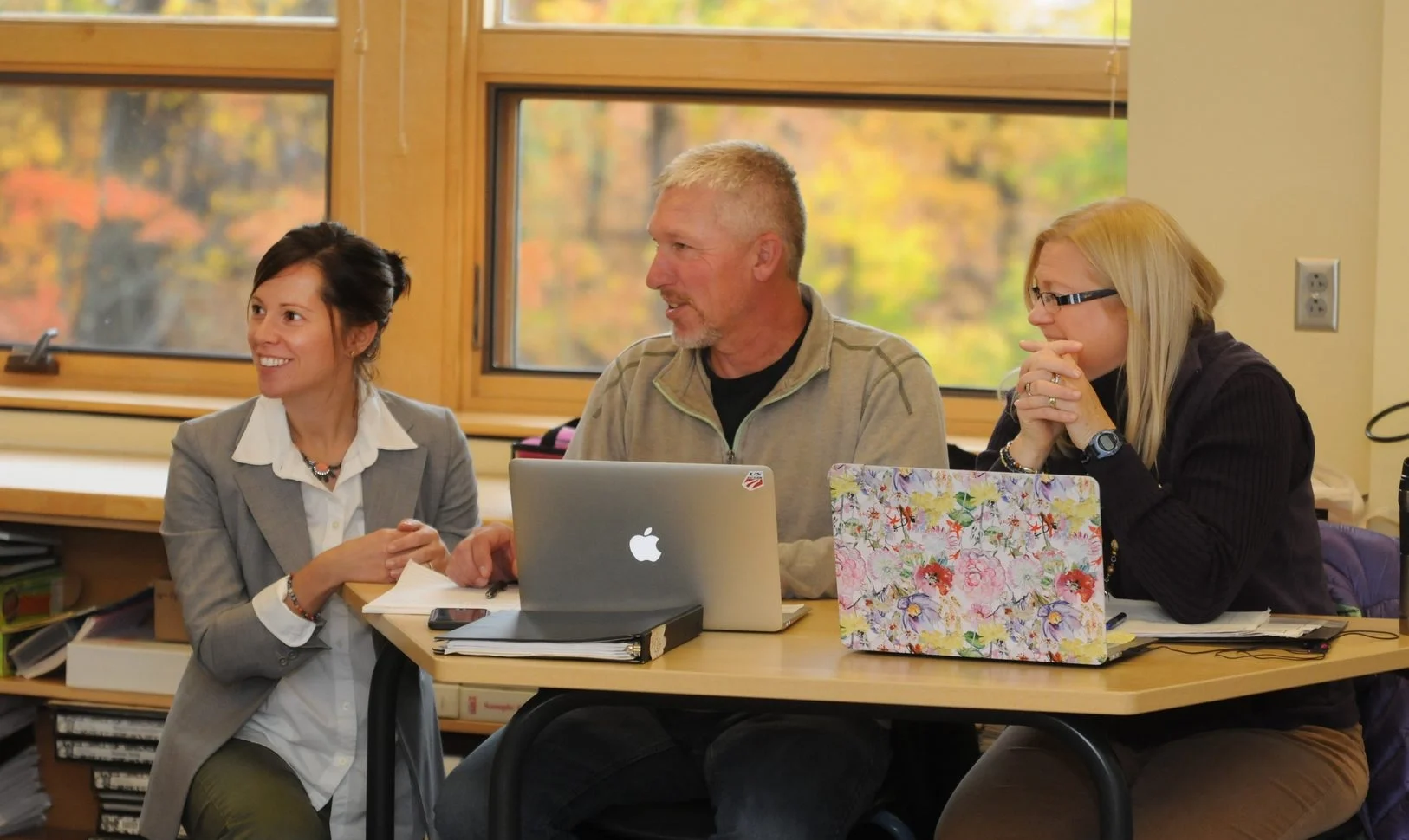 Three adults seated at a table in a classroom, discussing with laptops open in front of them.