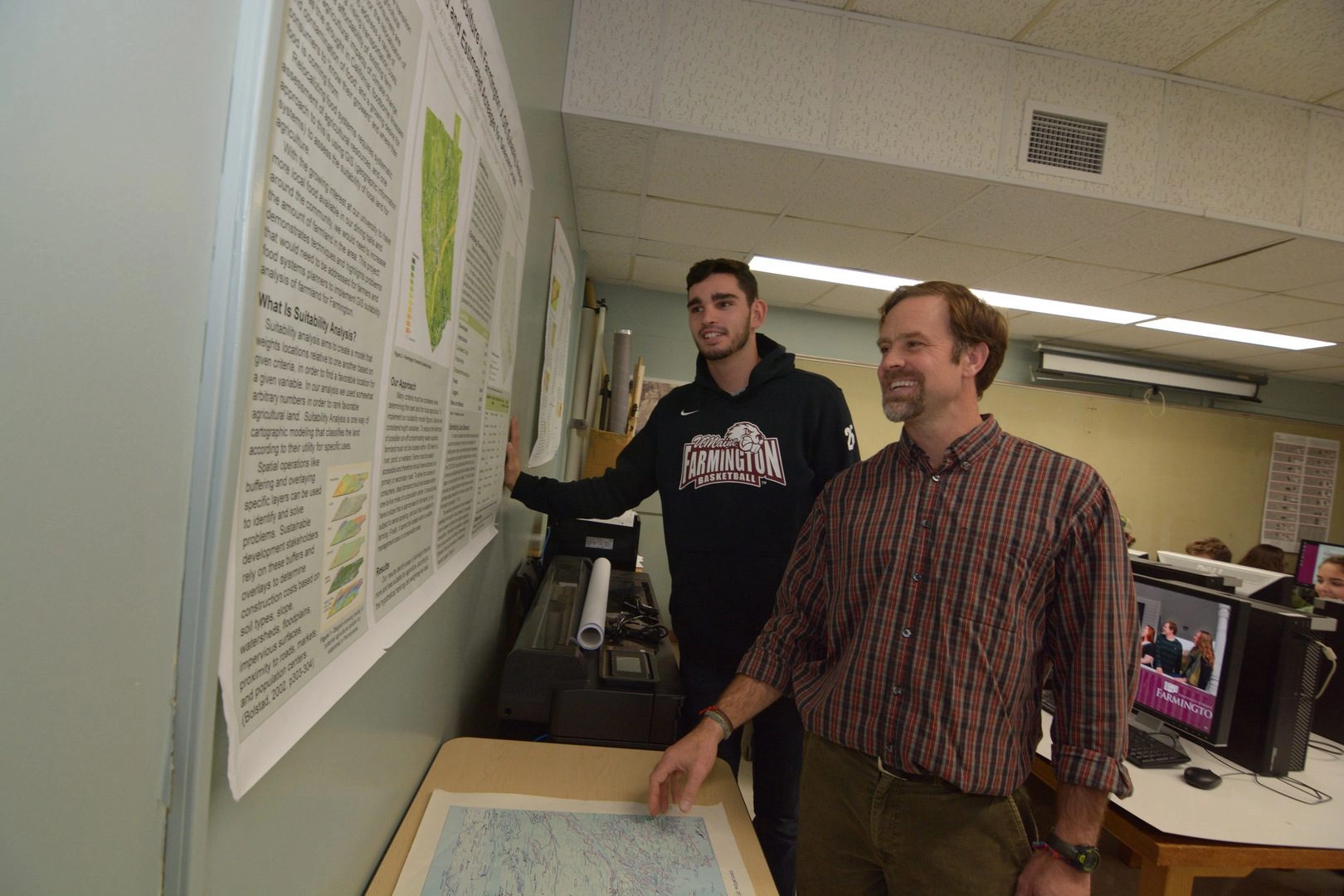 Two men stand at a wall of research posters, discussing the content and charts pinned up.