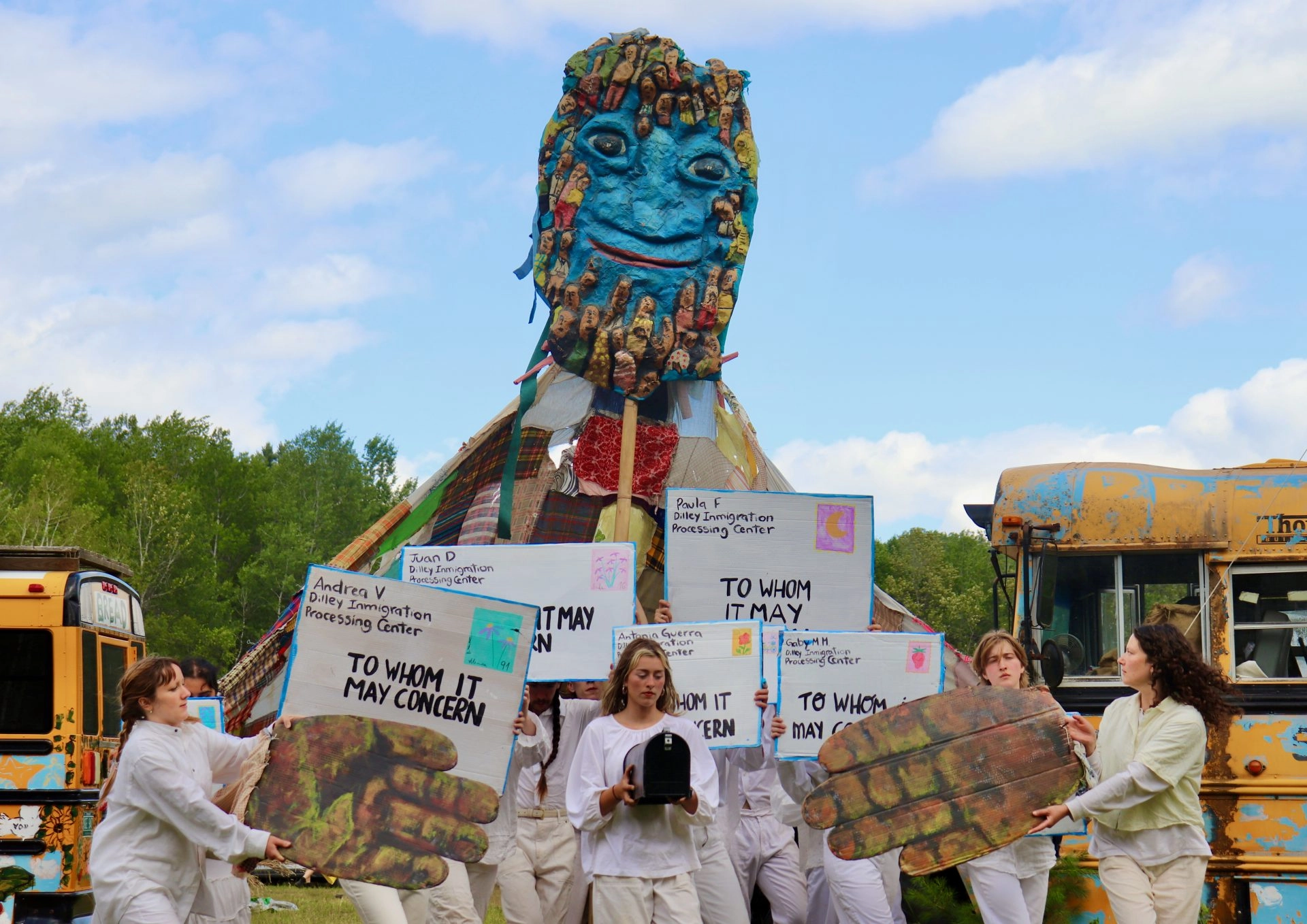 Group of people in white outfits holding signs about immigration processing in front of a tall blue sculpture head covered in small faces, outdoors.