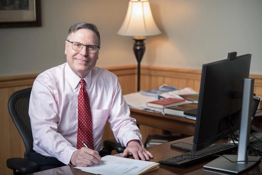 Smiling middle-aged man in a pink shirt and red tie signs a document at his desk, with a computer monitor and lamp in the background.