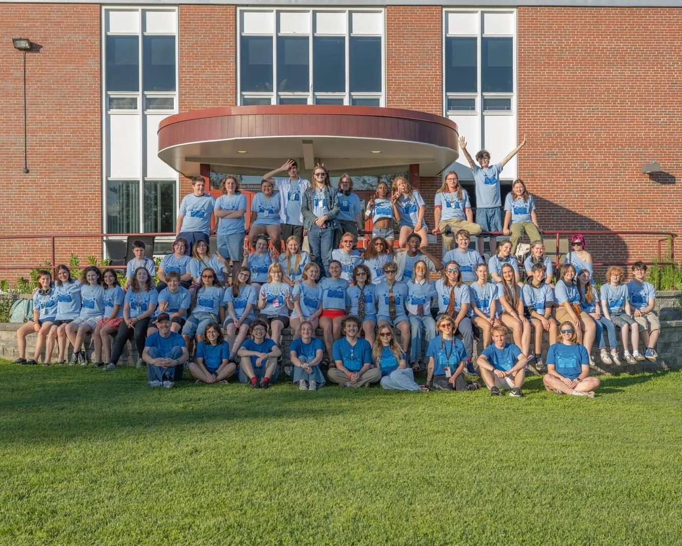 Large group of students in matching blue shirts posing on steps outside a brick school building. They are smiling and arranged in rows on the stairs and lawn.