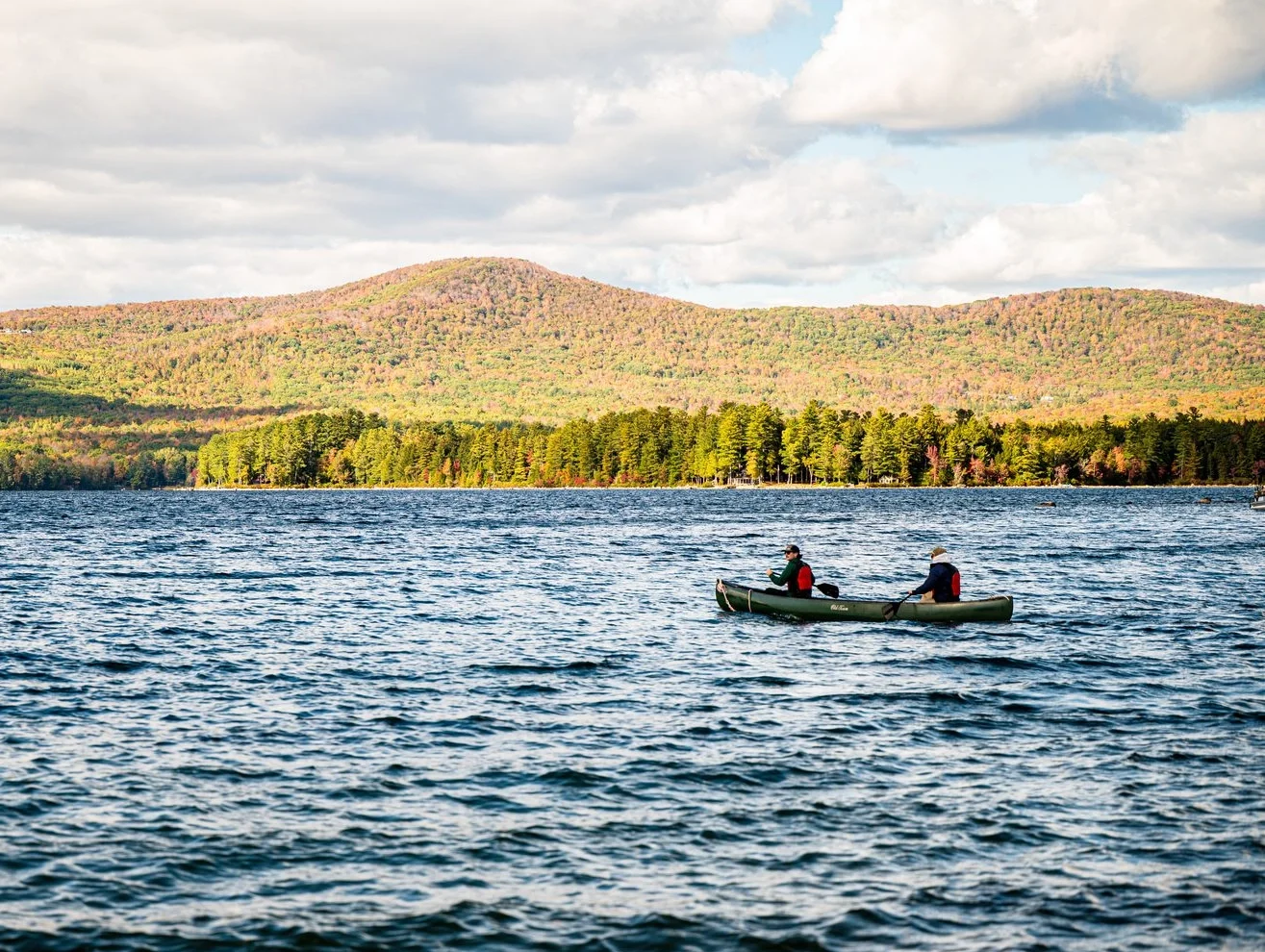 Two people in a canoe paddling on a blue lake with autumn-colored hills in the distance under a partly cloudy sky