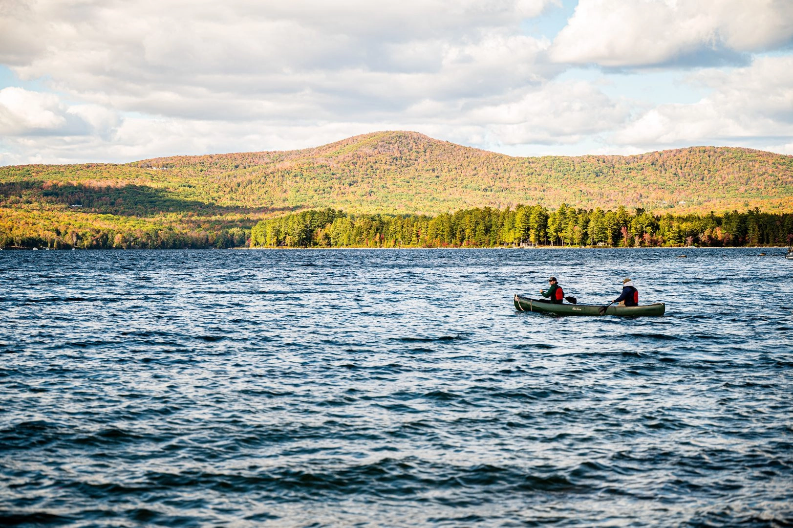 Two people in a canoe paddling on a blue lake with autumn-colored hills in the distance under a partly cloudy sky