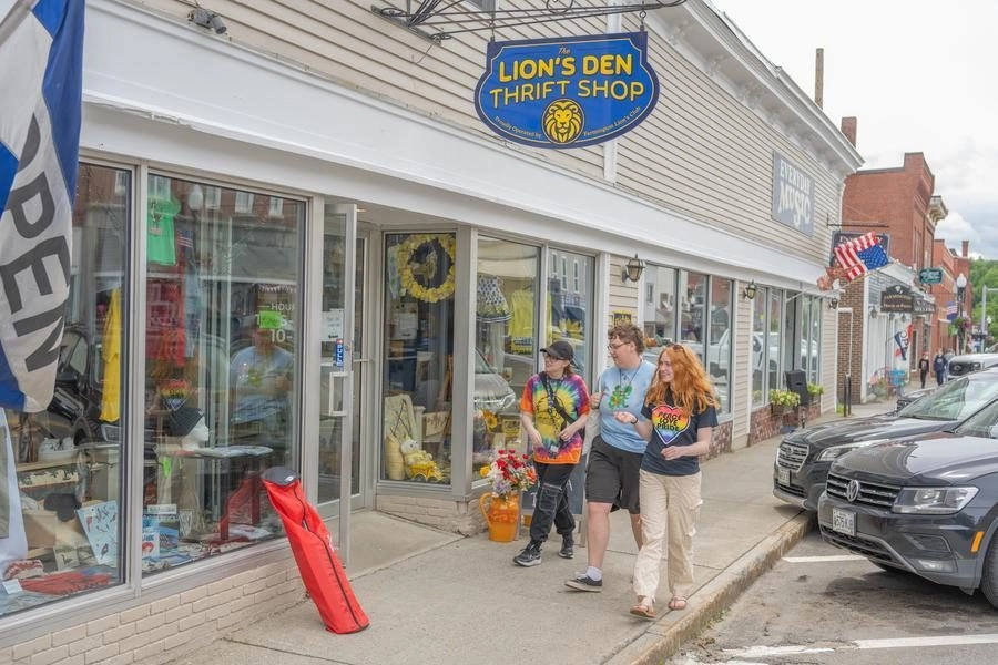 Three friends walk past The Lion's Den Thrift Shop on a sunny street, storefront windows displaying merchandise.