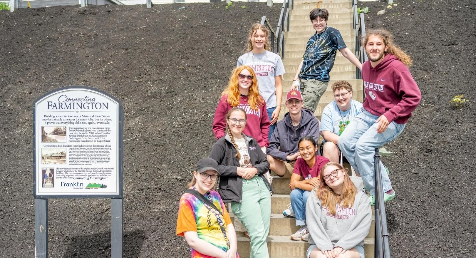 Group of students posed on an outdoor stairway beside a sign reading 'Connecting Farmington'.