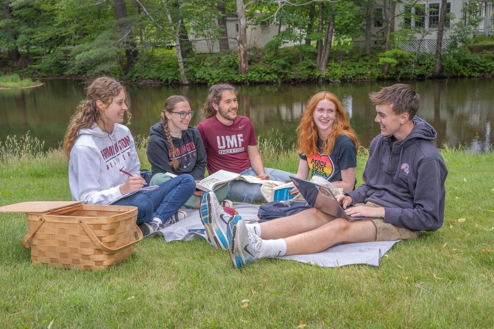 Students sitting on grass in front of pond studying on a picnic basket.