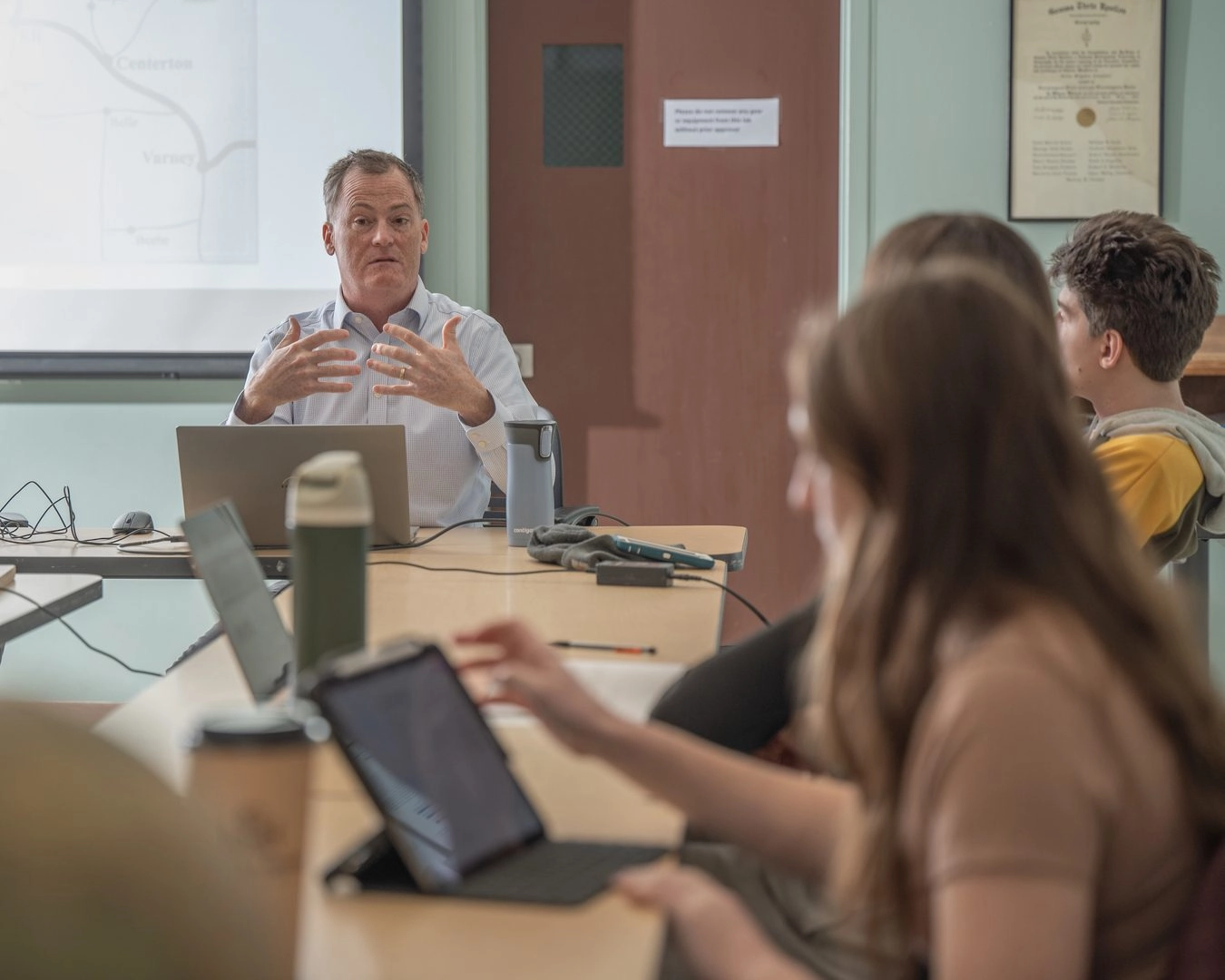 Instructor at head of table teaching a class with students using laptops and tablets in a classroom setting