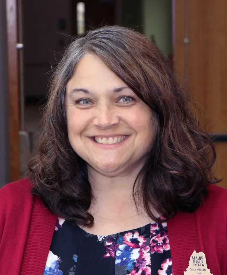 Portrait of a smiling woman with shoulder-length dark hair, wearing a red cardigan over a floral top, indoors; badge reads 'Maine Teacher of the Year'.
