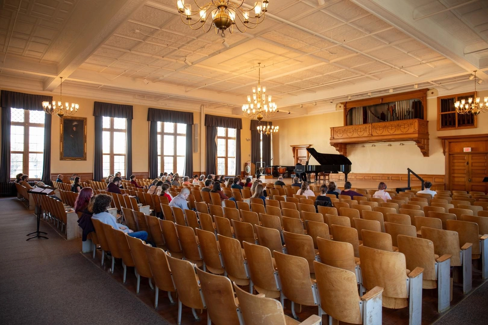Auditorium with wooden seats facing a grand piano onstage, chandeliers hanging from a high ceiling and tall windows along the walls.