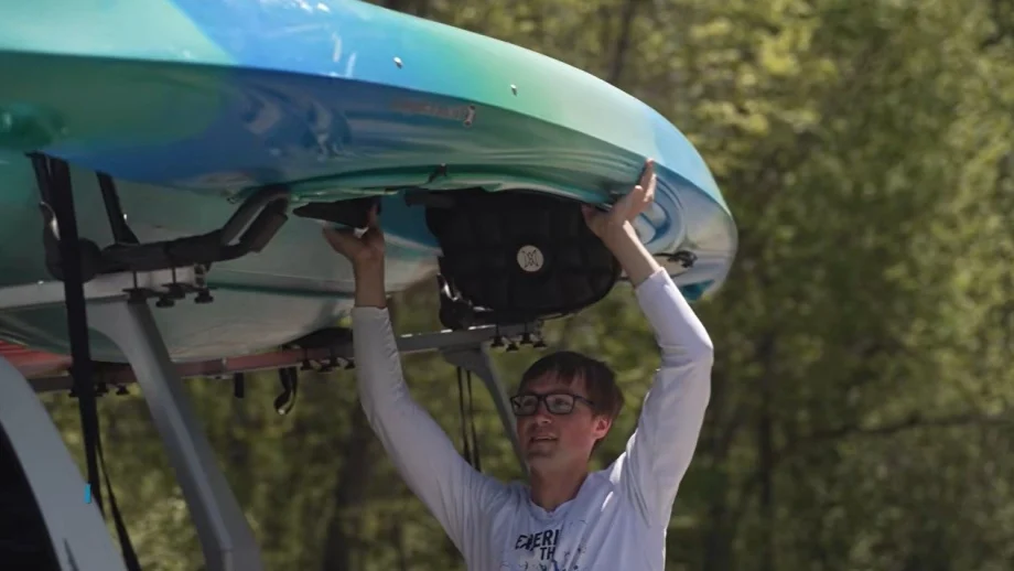 Man with glasses lifts a blue-green kayak onto a car roof rack outdoors.
