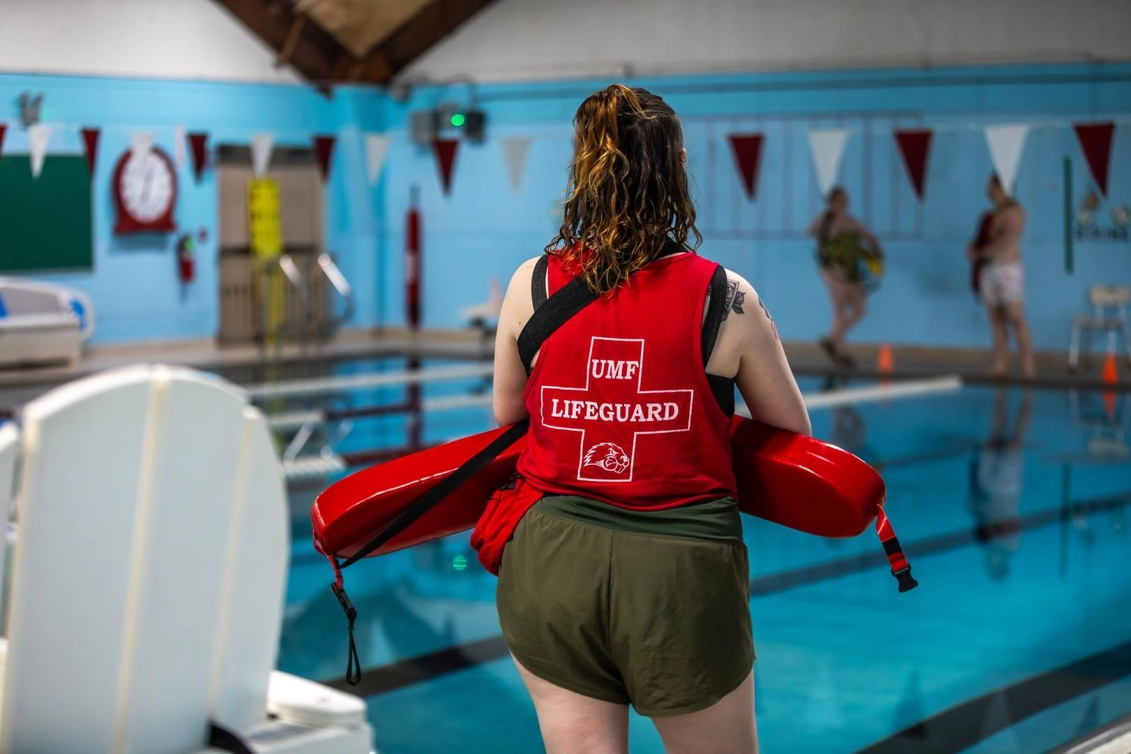 Back view of a lifeguard wearing a red UMF Lifeguard vest and green shorts, holding a red rescue tube by an indoor pool.