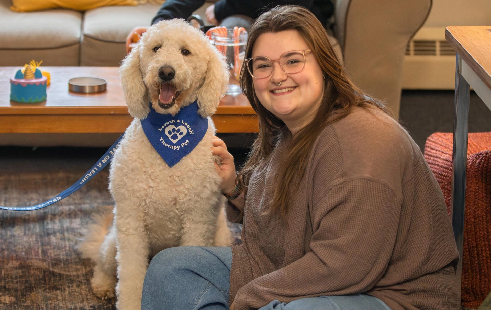 Smiling woman with glasses sits beside a fluffy white therapy dog wearing a blue bandana that reads 'Love on a Leash Therapy Pet'.