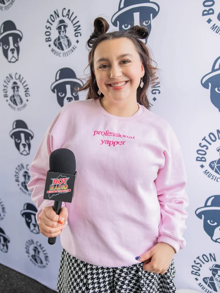 Smiling woman in a pink sweatshirt holding a microphone for an on-site interview at a Boston Calling backdrop festival.