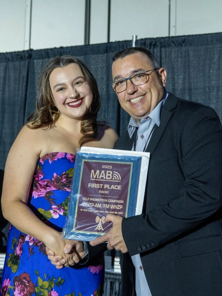 Smiling woman in a floral dress and man in a suit shake hands while holding a plaque that reads 'First Place' at an awards event.