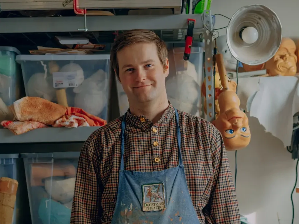 Young man in a plaid shirt and blue apron smiles at the camera in a cluttered workshop with plastic bins and a hanging mannequin head nearby.