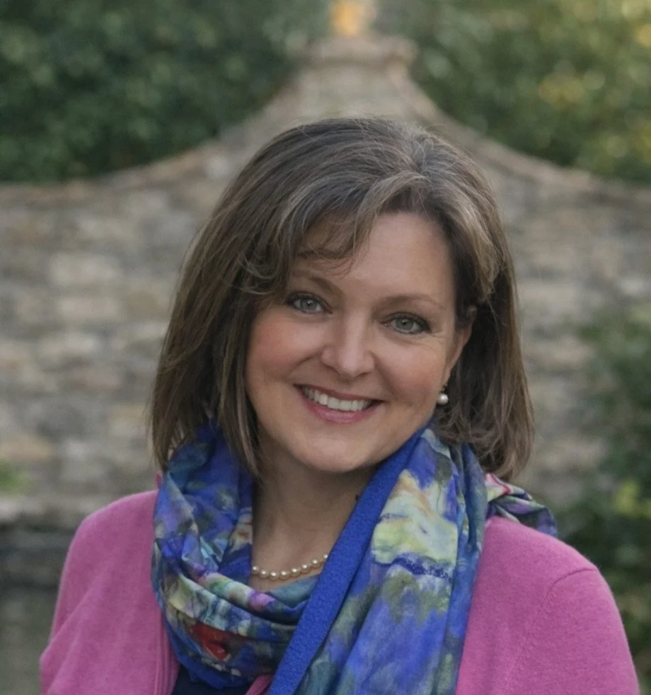 Smiling woman wearing a pink cardigan and a colorful scarf, outdoors near a stone wall.
