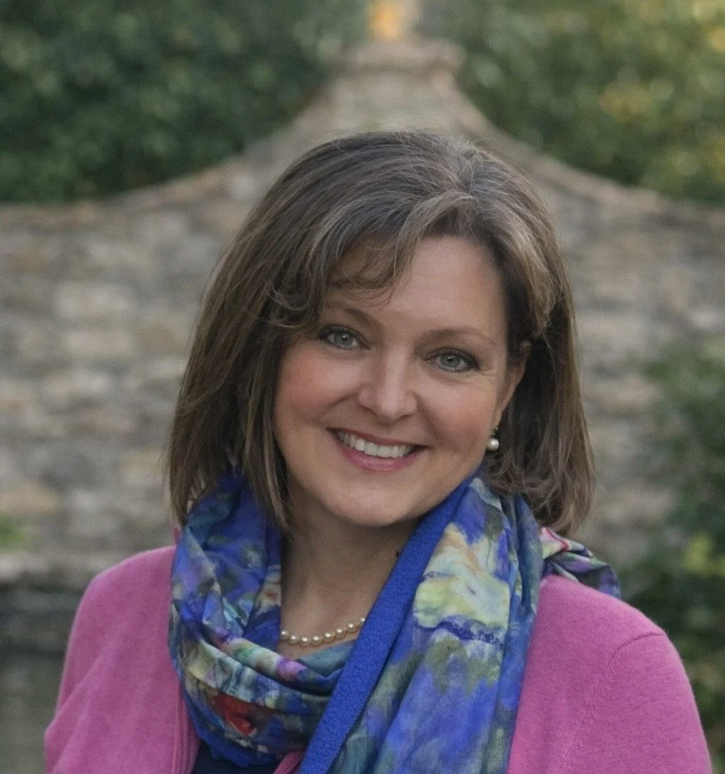 Smiling woman wearing a pink cardigan and a colorful scarf, outdoors near a stone wall.