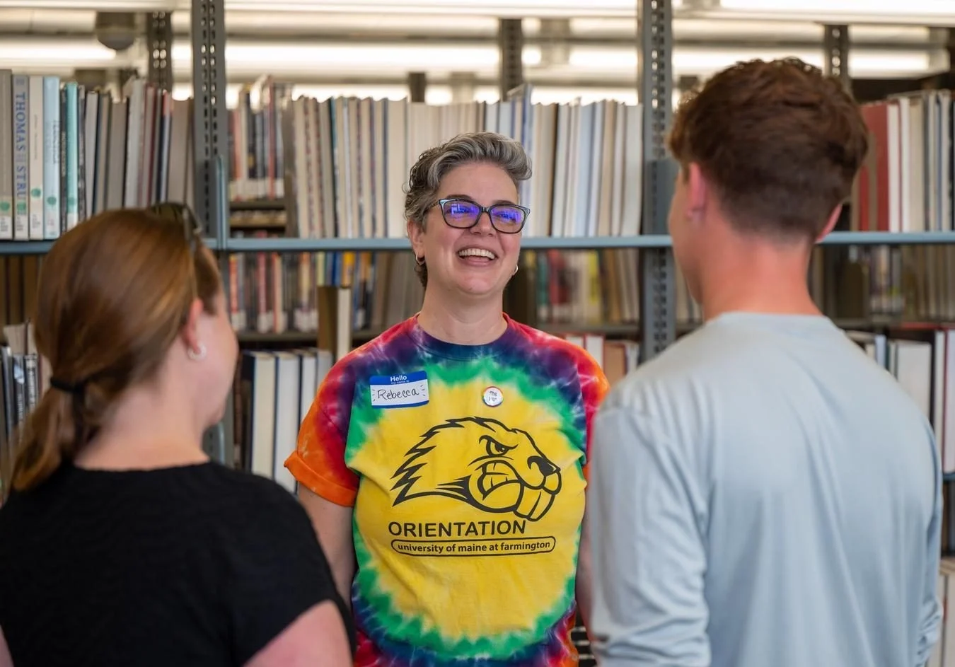 Smiling staff member Rebecca in a colorful tie-dye orientation shirt chats with two students in a library.