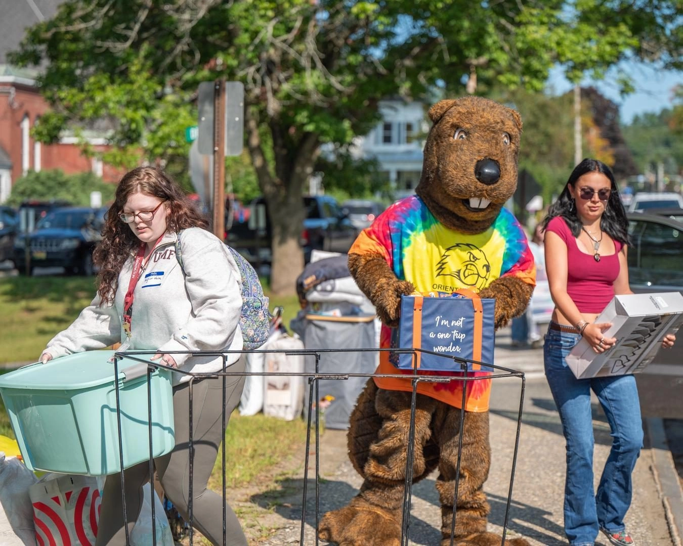 Bear mascot in a rainbow tie-dye shirt holds a blue tote that reads 'I'm not a one trip wonder' at an outdoor street event, with two women nearby and a shopping cart in the foreground.