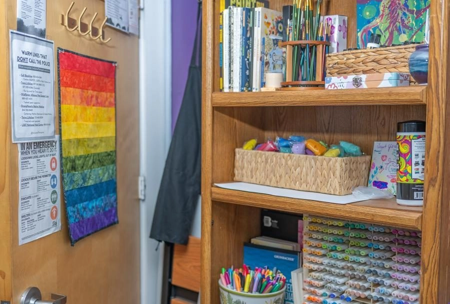 Open wooden shelf filled with art supplies—markers, colored pencils, paints, baskets, and notebooks; rainbow poster on the door nearby.