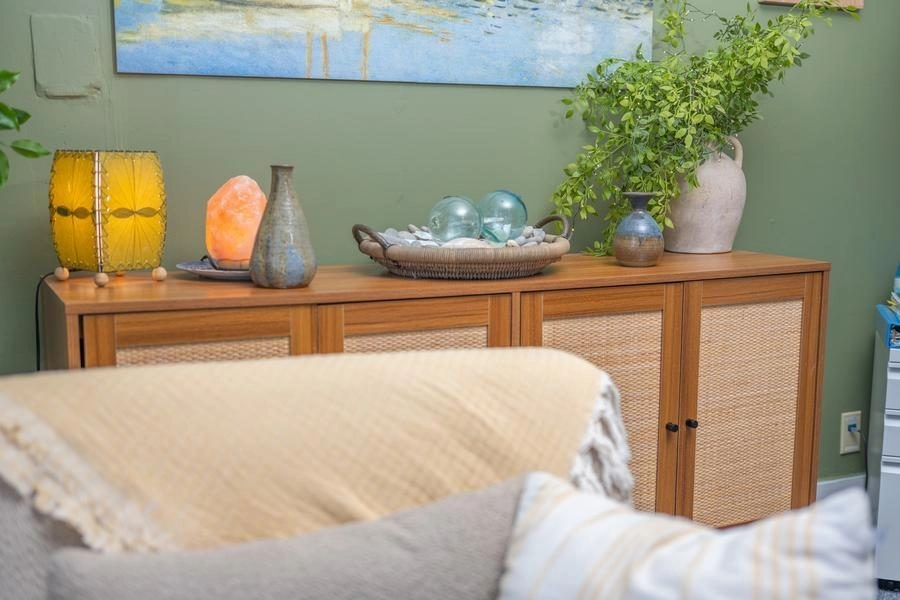 Wooden sideboard with woven doors, adorned by a yellow lamp, vases, and a plant against a green wall in a living room.