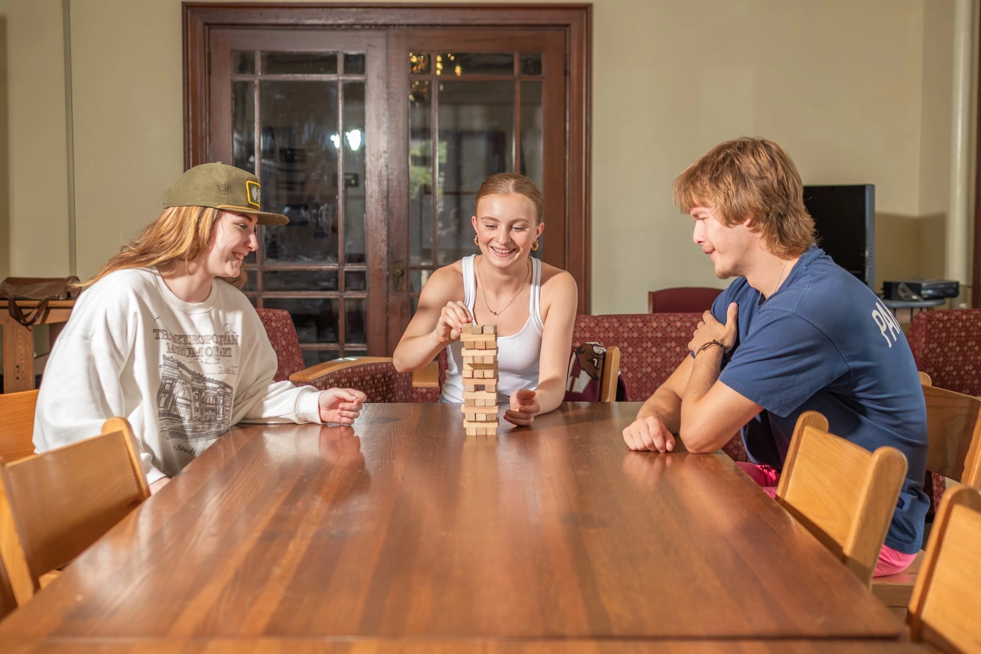 Three friends sitting at a wooden table playing Jenga and smiling.