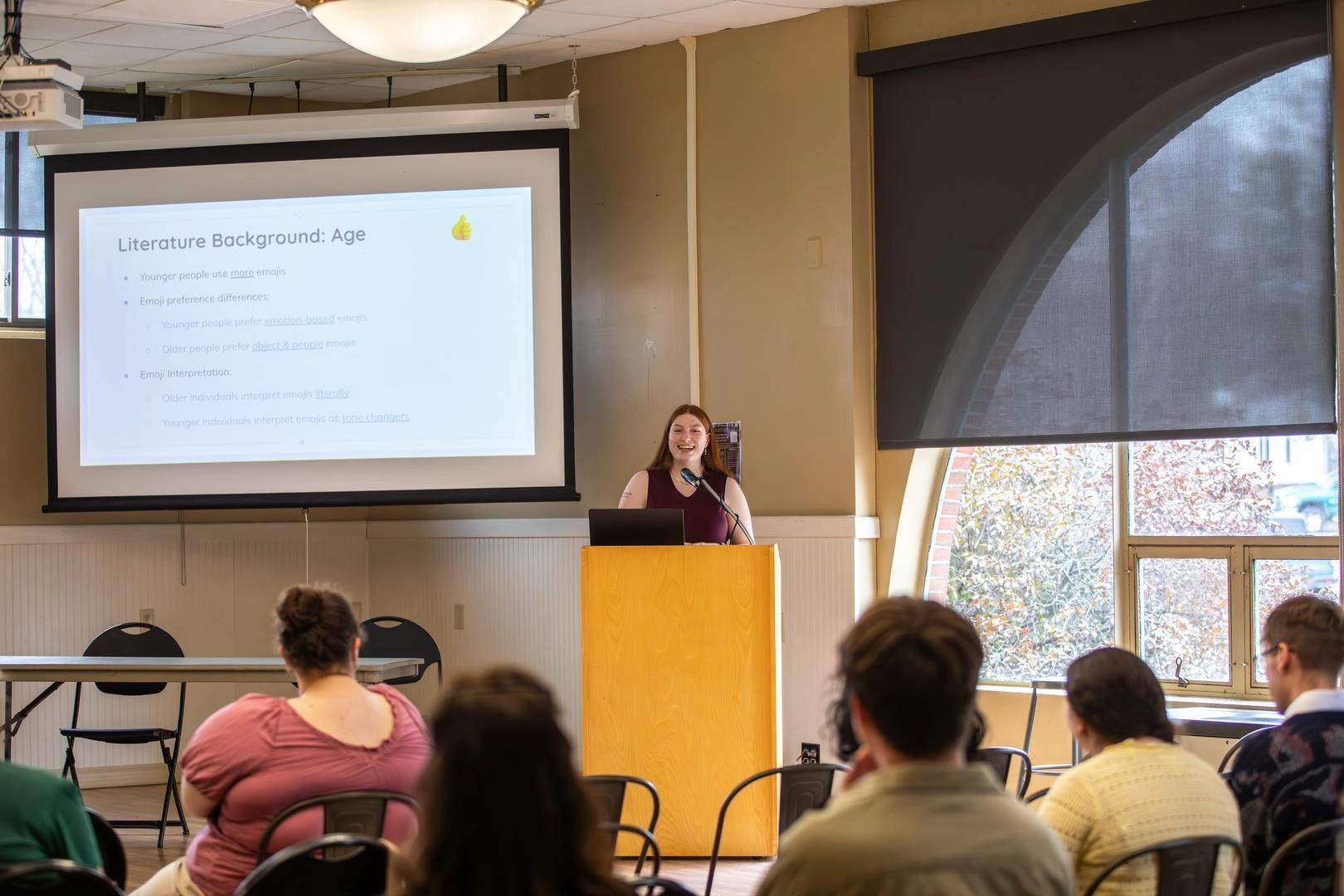 Woman presenting at a lectern in a classroom, slides titled 'Literature Background: Age' projected behind, audience seated.