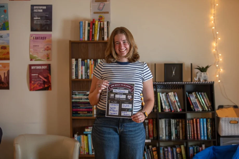 Smiling woman in a striped shirt holding a back-to-school poster in a cozy room with bookshelves and hanging fairy lights behind her.