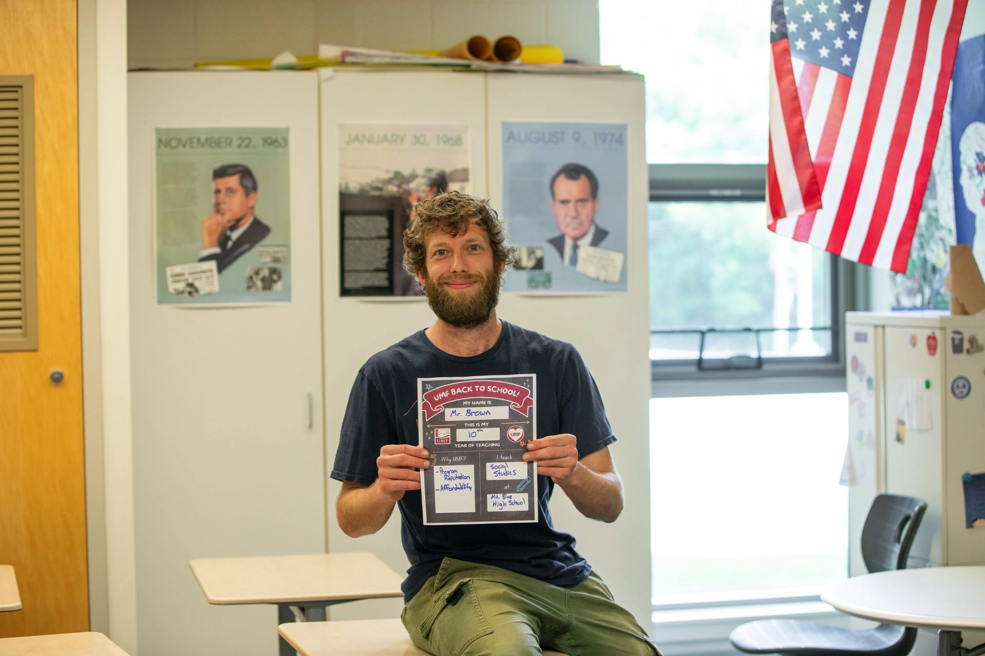 Bearded man in a navy shirt sits on a desk in a classroom, holding a large illustrated poster about teaching. A U.S. flag hangs by windows behind him.