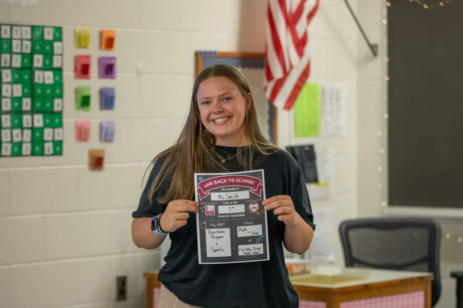 Smiling girl in a classroom holding a back-to-school poster with her name on it (Ms. Smith).