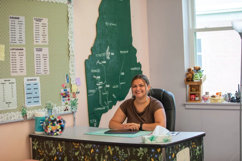 Smiling woman seated at a desk in an office/classroom, with a large green map on the wall and schedules on a corkboard behind her.