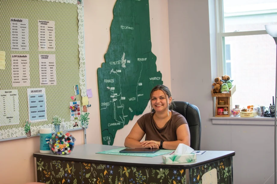Smiling woman seated at a desk in an classroom, with a large green map of Maine on the wall and schedules on a corkboard behind her.