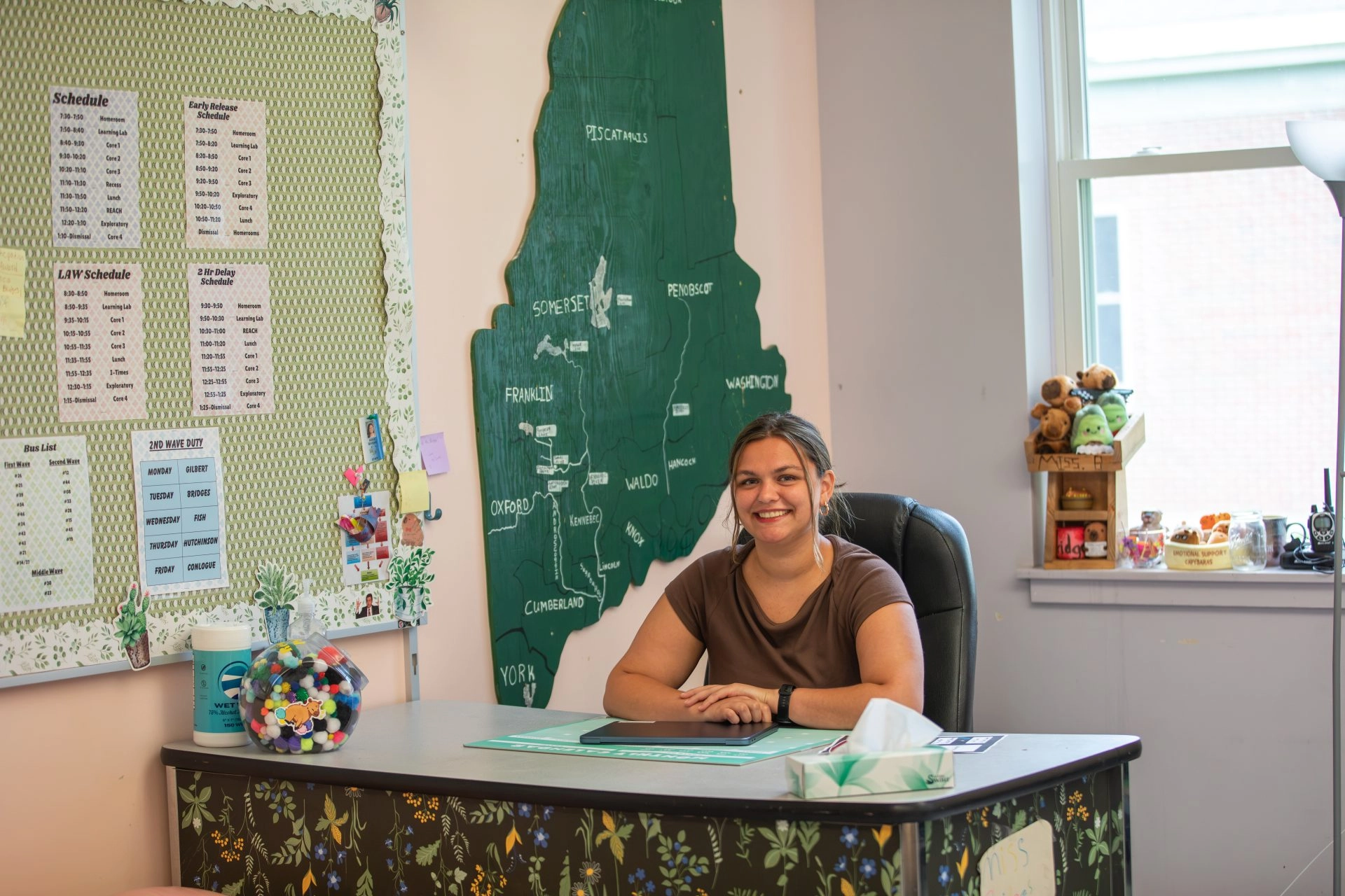 Smiling woman seated at a desk in an classroom, with a large green map of Maine on the wall and schedules on a corkboard behind her.
