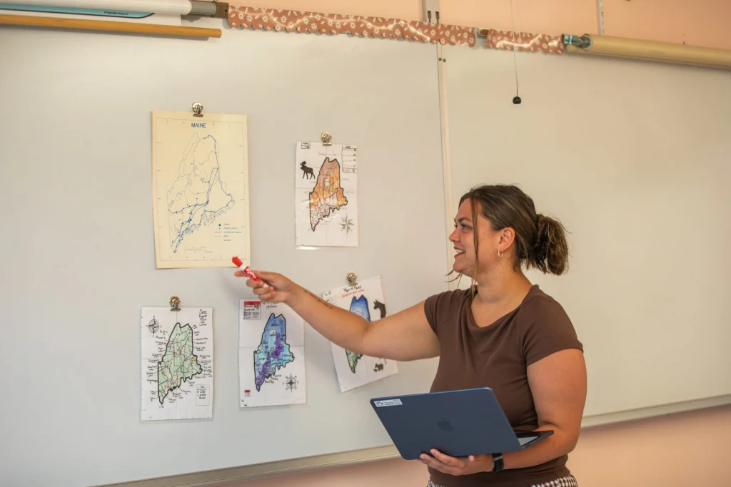 Woman in a brown shirt points at maps clipped to a classroom wall, holding a red marker and a blue laptop in front of a whiteboard.